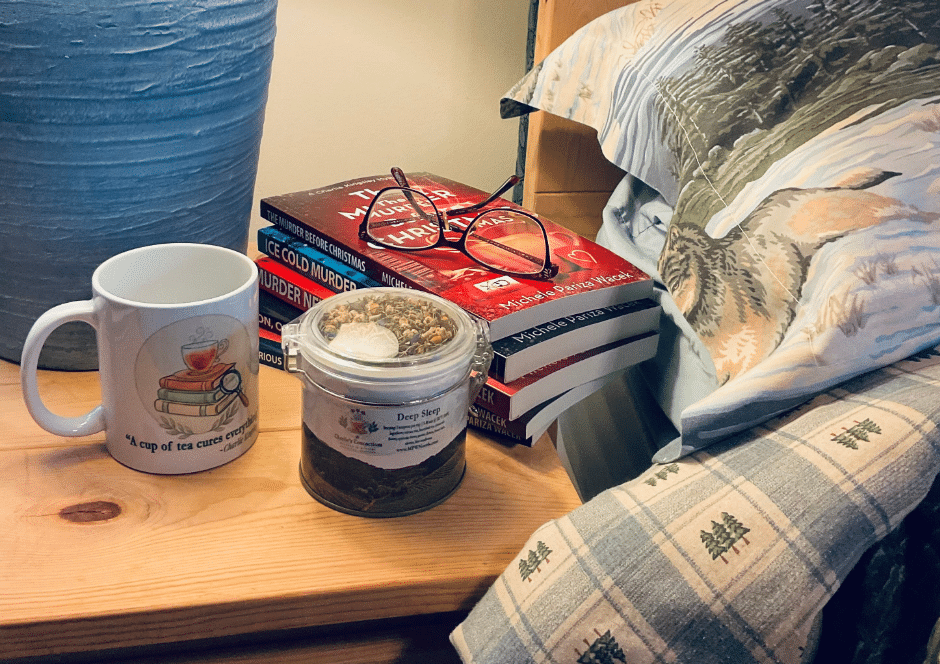 Charlie's Concoctions Deep Sleep tea tin and "A cup of tea cures everything" mug on a bedside table, with a stack of Charlie Kingsley Mystery books by Michele Pariza Wacek and reading glasses nearby